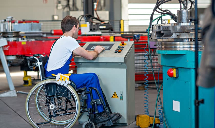 Hombre en silla de ruedas operando una máquina industrial desde un panel de control dentro de una fábrica. Lleva puesto un mono de trabajo azul y una camiseta blanca, y está concentrado en la pantalla del panel. El entorno es claramente industrial, con maquinaria pesada alrededor.